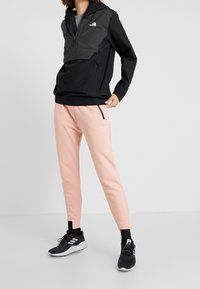 Schwarze leichte Kapuzenjacke mit einer Vordertasche, kombiniert mit pinkfarbenen schmal zulaufenden Sweatpants und schwarzen Sportschuhen. Schlichtes, funktionales Design.