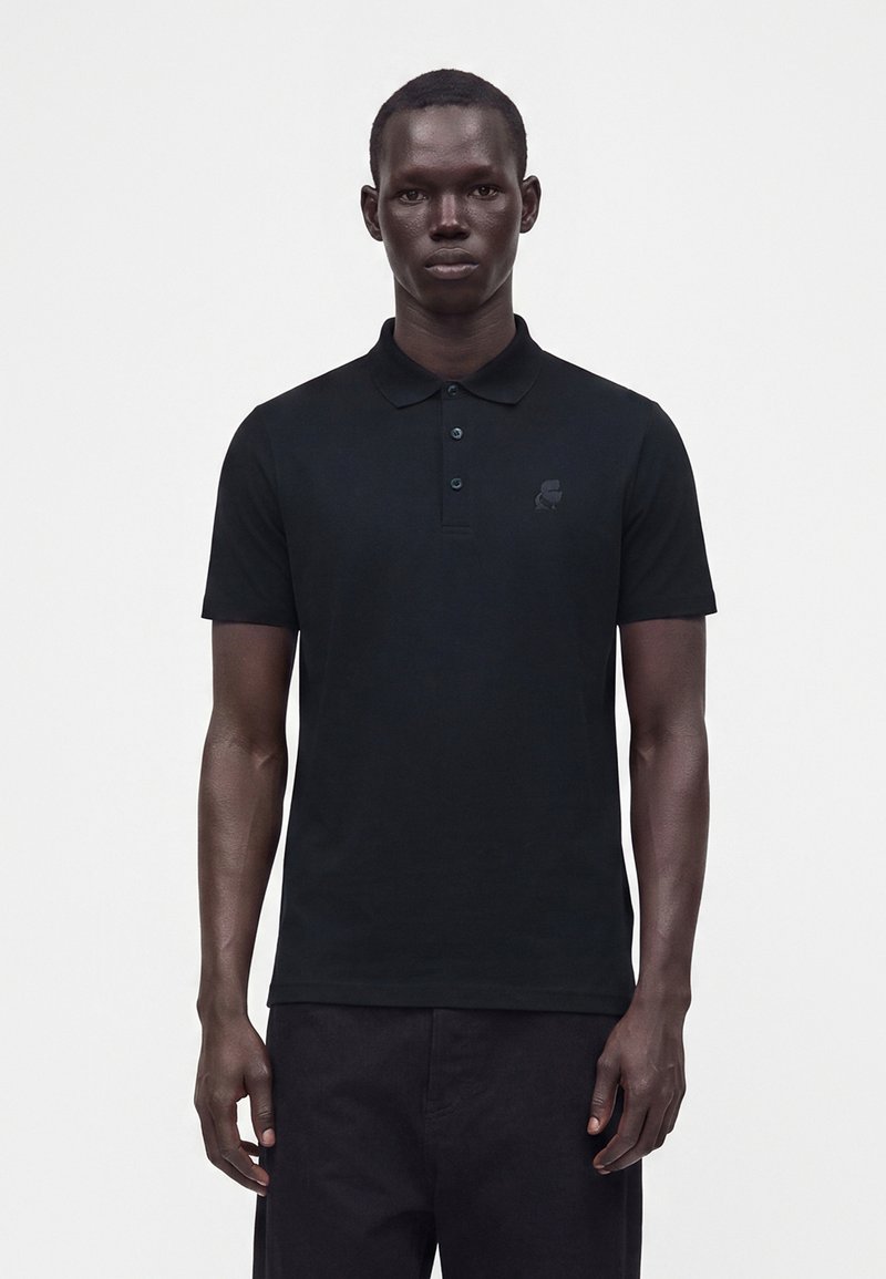 Homme à la peau foncée portant un polo noir à manches courtes et un pantalon noir, debout devant un fond clair uni.