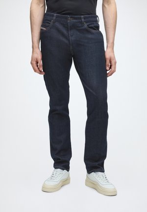Muž nosi tmavomodré slim-fit džínsy a biele tenisky, stojí pred jednoliatym svetlým pozadím.