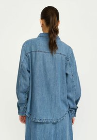 Denim brīvā stila krekls ar garām piedurknēm, zilā krāsā, ar pogu aizdari priekšpusē un izliekto apakšmalu. Šuvju un aproču dekoratīvās šuvju detaļas.