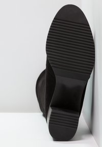 Bottine en daim noir avec une semelle en caoutchouc plate. La semelle comporte des rainures horizontales pour l'adhérence, et le talon présente un léger rehaussement.