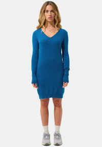 Donna con capelli biondi ondulati, indossa un abito di maglia lungo blu, calze bianche, scarpe grigie e una collana d'oro, mentre è in piedi davanti a uno sfondo bianco.