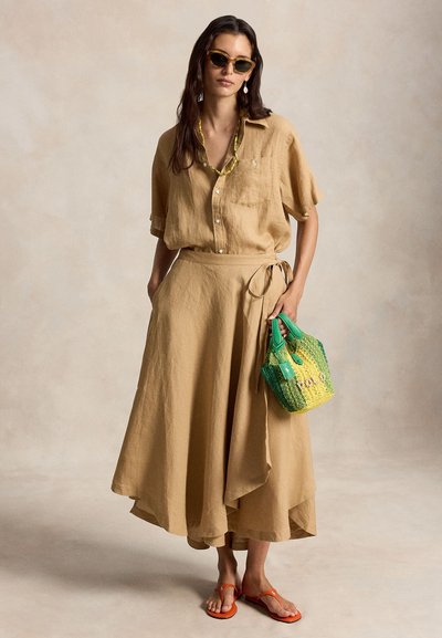 Moteris dėvi gelsvos spalvos lino marškinius ir apsiaustą sijoną, akinius nuo saulės, geltoną vėrinį, laiko žalią ir geltoną pintą krepšį bei dėvi oranžines basutes.