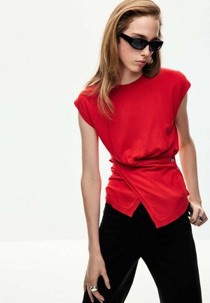 Mujer con cabello largo castaño claro con gafas de sol negras, camiseta roja sin mangas con botones y pantalones negros sobre un fondo claro y sencillo.
