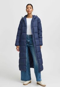 Námornícky puffer kabát s dlhým prešívaným dizajnom, kapucňou a predným zipsom. Modelka ho kombinuje s bielym topom a nohavicami širokého strihu.