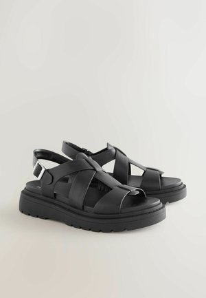 Schwarze Ledersandalen mit dicker Sohle, ausgestattet mit überkreuzten Riemen und verstellbarer Schnalle. Glatte Textur und offenes Fersendesign.