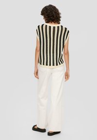 Kobieta o kręconych włosach, ubrana w bezrękawnik z dzianiny w czarno-kremowe paski, białe spodnie o szerokich nogawkach i czarne sandały, stoi tyłem.