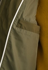 Jachetă verde măsliniu, cu un material neted, fermoar alb în contrast și un buzunar pe piept. Fundalul prezintă o țesătură texturată de muștar.