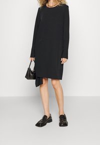 Musta pitkähihainen mekko, jossa on rento siluetti, kaarrettu helma ja pehmeä kangas. Yhdistetty kiiltäviin mustiin loafereihin ja pieneen laukkuun.