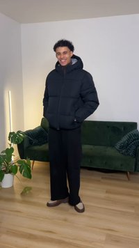 Črni puhasti plašč s kapuco, širokim krojem in žepki na straneh. Kombiniran z širokimi črnimi hlačami in rjavimi mokasini. Ozadje z zelenim kavčem.