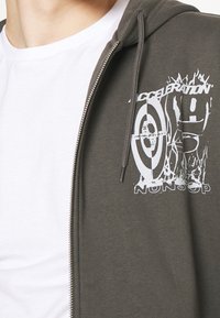 Primer plano de una persona que lleva una camiseta blanca y una sudadera oliva verde con la cremallera abierta, con el texto gráfico blanco "Aceleración Infinita" y un diseño de diana.