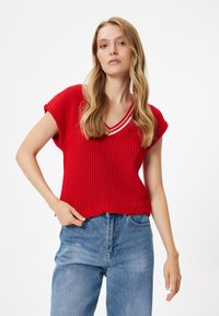 Jersey de punto rojo con mangas cortas, escote en forma de V y acentos de franjas blancas en el cuello. Combinado con jeans de talle alto azul.