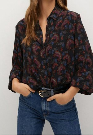 Femme portant un chemisier noir à manches longues avec un imprimé paisley rouge et bleu, rentré dans un jean bleu avec une ceinture noire, mains dans les poches.