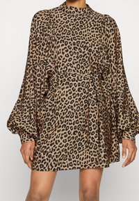 Šaty s leopardím potiskem z měkké tkaniny, vysoký límec, balónové rukávy a přiléhavý pas se stahovací šňůrou; neutrální tóny s černými skvrnami.