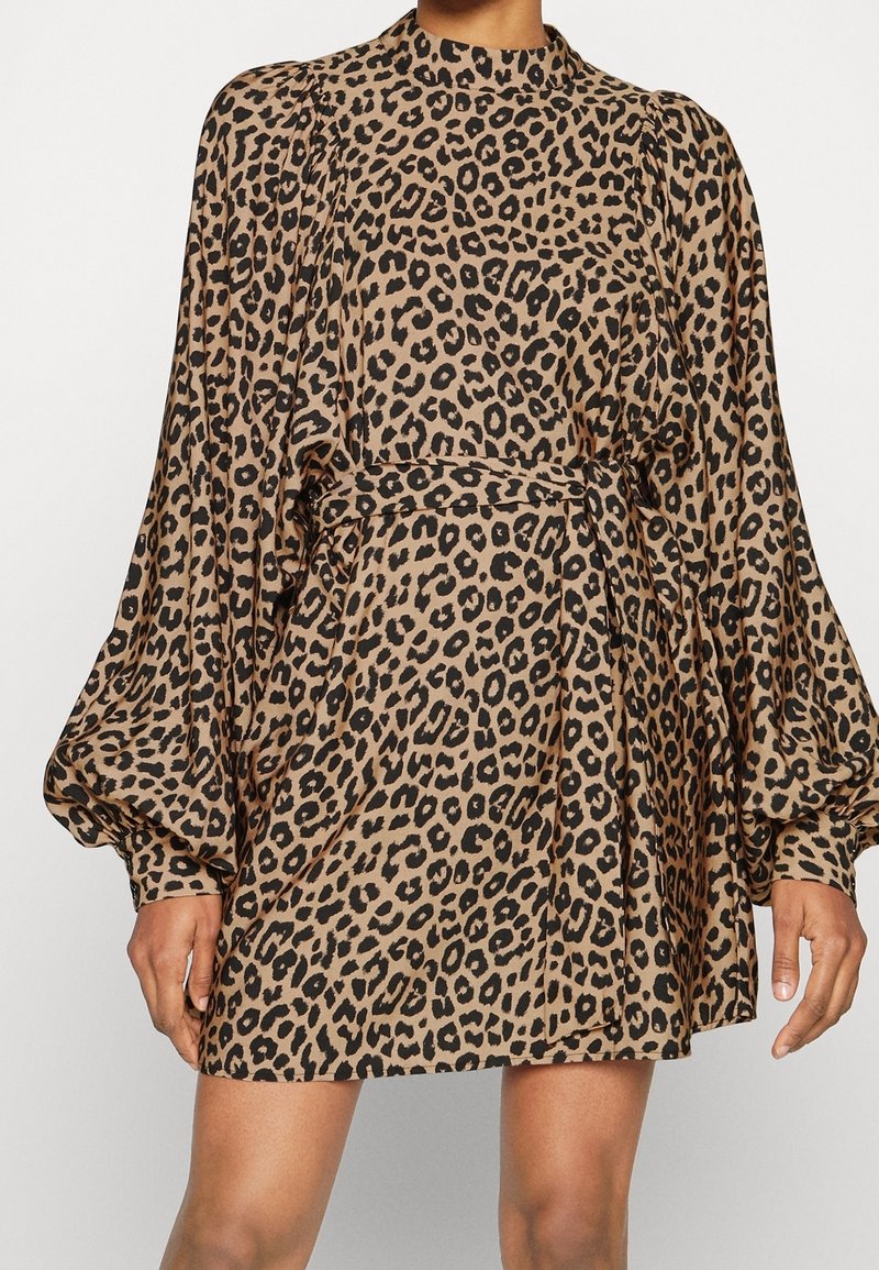 Šaty s leopardím potiskem z měkké tkaniny, vysoký límec, balónové rukávy a přiléhavý pas se stahovací šňůrou; neutrální tóny s černými skvrnami.