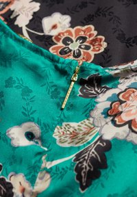 Tkanina w kwiatowy wzór pokazuje żywą zieloną sekcję o gładkiej teksturze, z złotym zamkiem błyskawicznym oznaczonym napisem "SUPERDRY" na szwie.