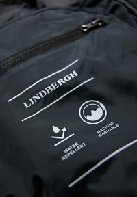 Svart tyg med vit tryckt text och ikoner som indikerar "LINDBERGH", "vattenavvisande" och "maskintvättbar." Har ett blixtlåsdetalj.