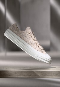 Jasno beżowy sneaker z teksturowanym wzorem sznurowadeł, białą gumową podeszwą i płaskimi sznurowadłami, zawieszony na miękkim szarym tle.