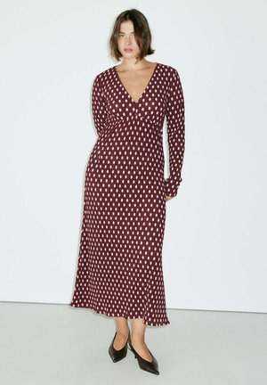 Femme debout portant une robe bordeaux à manches longues avec des pois blancs et des chaussures noires à bouts pointus et talons bas sur un fond uni.