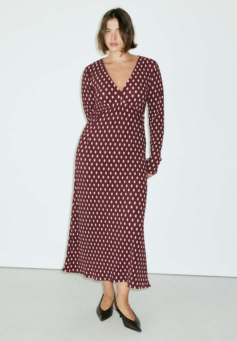 Femme debout portant une robe bordeaux à manches longues avec des pois blancs et des chaussures noires à bouts pointus et talons bas sur un fond uni.