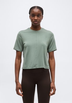 Woman with braided hair wearing a light green short-sleeve shirt and black leggings, standing against a plain white background.