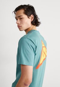 Camiseta de algodón turquesa de manga corta, con un gran diseño geométrico en naranja y amarillo en la parte posterior. Textura suave, cuello redondo.