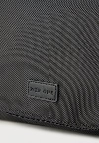 Μαύρη υφασμάτινη τσάντα με υφή στην επιφάνεια, ορθογώνιο σχήμα και δερμάτινη ετικέτα με ανάγλυφη την επιγραφή "PIER ONE." Ορατή λεπτομέρεια ραφής κατά μήκος των ακρών.