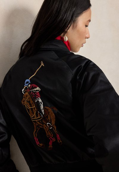 Frau mit dunklen Haaren, die eine schwarze Jacke trägt, auf der ein bestickter Polospieler zu sehen ist, der auf einem Pferd mit einem Schlägel einen Ball schlägt.