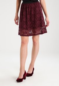 Jupe en dentelle bordeaux avec une taille élastique, dotée de motifs floraux et d'un bord inférieur semi-transparent, associée à des talons en velours rouge.