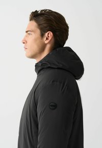 Schwarze, kapuzenjacke mit glatter Textur. Verfügt über ein rundes schwarzes Logo auf dem linken Ärmel und ein elegantes Design.