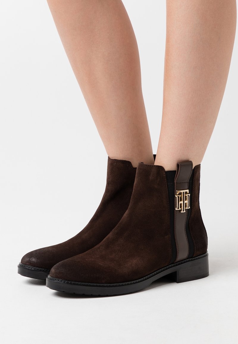 Bottes courtes en daim brun foncé avec des panneaux latéraux élastiques noirs et un emblème de logo doré, portées sur des jambes nues sur fond blanc.