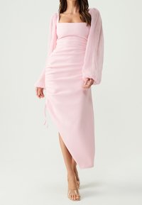 Woman wearing a light pink ruched midi dress with puffed long sleeves and square neckline, paired with clear open-toe heels.
