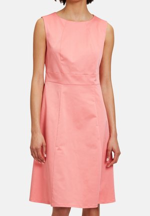 Vrouw draagt een mouwloze, knielange roze jurk met een getailleerde taille en een eenvoudige ronde halslijn tegen een effen achtergrond.