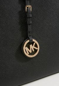 Czarna torebka ze skóry o fakturze z złotym okrągłym logo Michael Kors zawieszonym na regulowanym pasku.