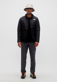 Άνδρας που φοράει μαύρο μπουφάν με φουσκωτή επένδυση, σκούρα γκρι παντελόνια, μπεζ καπέλο τύπου bucket, γυαλιά ηλίου και παπούτσια για τρέξιμο σε μονοπάτια με κόκκινες κορδόνια.