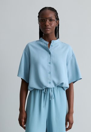 Vrouw met gevlochten haar, grote bril en een losse lichtblauwe blouse met bijpassende broek met trekkoord, tegen een effen grijze achtergrond.