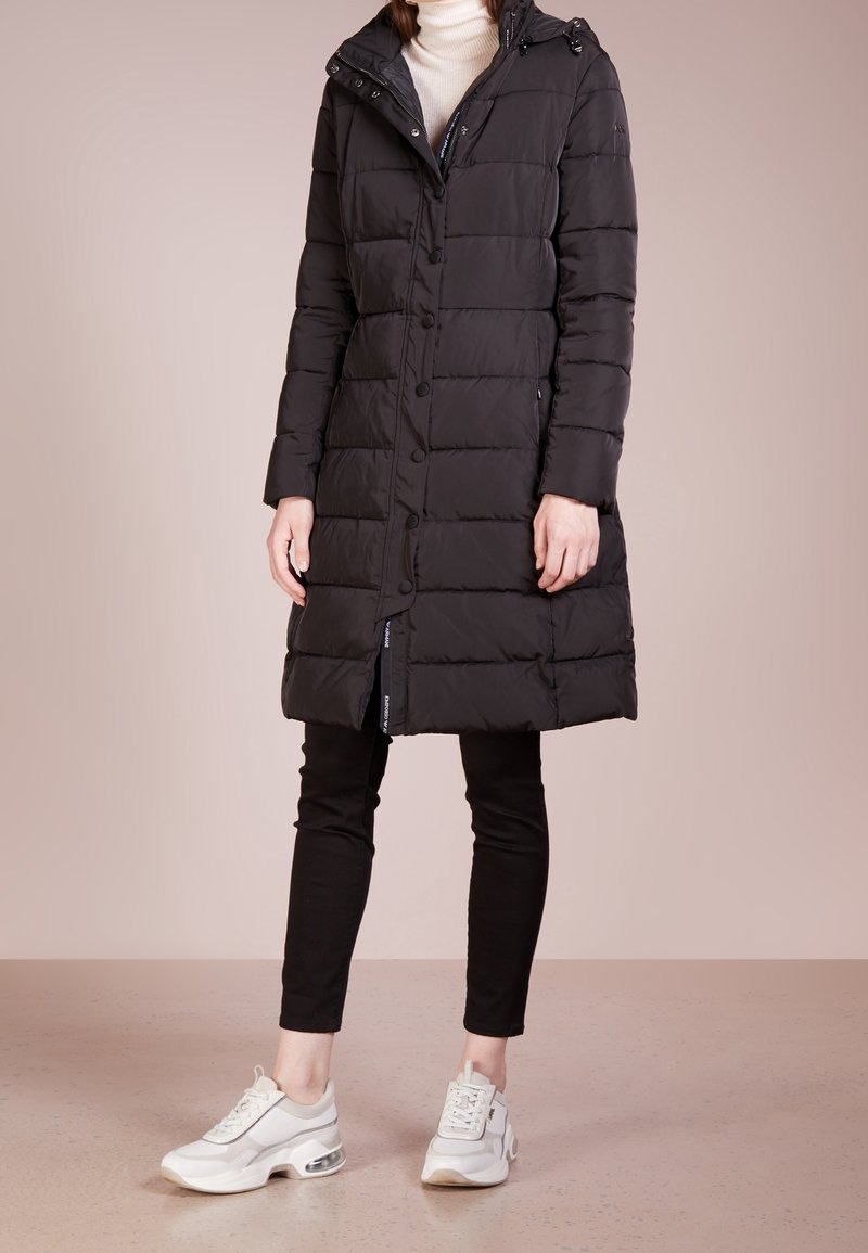 Schwarze Pufferjacke mit Knopfleiste und Kapuze, mit einem gesteppte Design und Seitentaschen. Getragen mit schwarzen Hosen und weißen Sneakers.