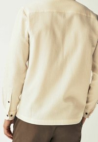 Chemise texturée beige clair avec un motif carreaux subtil, poignets à boutons et ourlet droit. Le matériau semble léger et respirant.