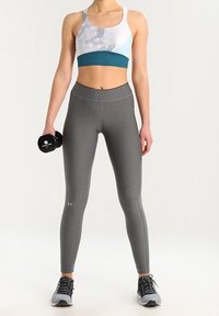 Stanik sportowy w bieli i zieleni w geometryczne wzory, w połączeniu z szarymi legginsami o wysokim stanie. Modelka trzyma czarny hantel.
