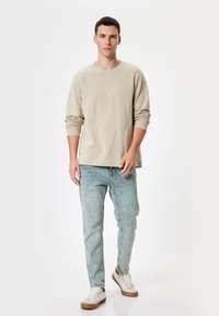 Camisa de manga larga beige, jeans azul claros con acabado desgastado y zapatillas blancas con detalles en marrón. Ajuste sencillo y relajado.