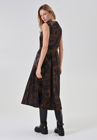 Blumenmusterkleid in Schwarz mit ärmellosem Design, figurnahem Oberteil und schwingendem Rock. Kombiniert mit knielangen schwarzen Stiefeln.