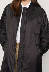 Černá bomber bunda s texturovanou podšívkou a žebrovanými manžetami. Má přední zip a boční kapsy. Nositá přes bílou košili.