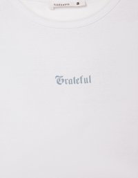 T-shirt bianco in cotone con testo azzurro chiaro che recita "Grato" in un carattere corsivo. Scollo rotondo semplice e texture morbida.