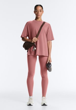 Femeie în picioare, purtând un tricou supradimensionat de culoare roz prăfuit și leggings, pantofi sport albi, având o geantă purtată în diagonală și ținând un termos de călătorie negru.