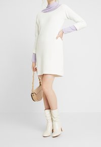 Donna che indossa un vestito di maglia bianco sopra maniche lunghe lavanda, tiene una borsa beige con tracolla intrecciata e indossa stivaletti bianchi.