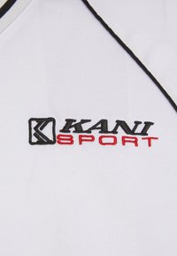 Balta sportinė marškinė su juodu ir raudonu siuvinėtu „KANI SPORT“ logotipu. Savybės - tekstūruota medžiaga ir kontrastingos juodos apdailos. 
