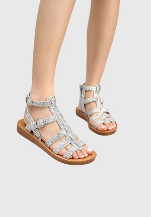 Jambes portant des sandales argentées pailletées à brides avec boucles, debout devant un fond gris clair.