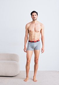 Szürke pamut boxer alsónadrág fekete és piros derékpánttal, rajta a "PUMA" logóval. A modell mezítláb áll, előre nézve egy minimális környezetben.