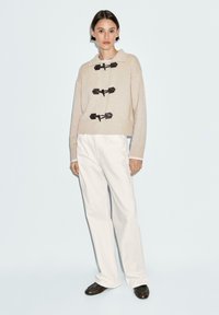 Beige knitted sweater with three black toggle closures, ribbed cuffs, and a wide collar, paired with relaxed white pants and dark shoes.