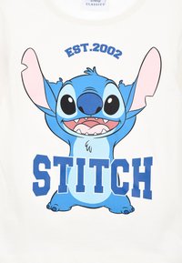 Grafisches T-Shirt mit einem blauen Cartoon-Charakter mit großen Ohren und ausdrucksvollen Augen. Der Text liest "EST.2002" und "STITCH" in fetten blauen Buchstaben.